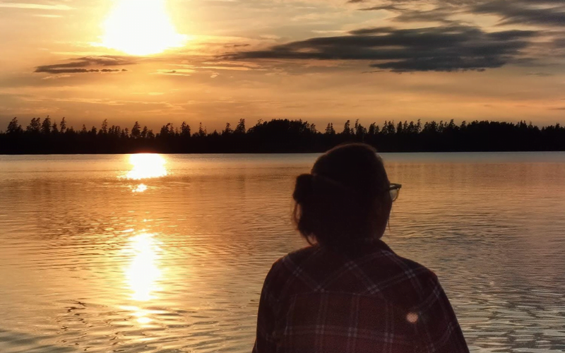 Foto van een vrouw die zit met haar rug richting de camera terwijl ze kijkt richting een meer met tijdens de ondergaande zon